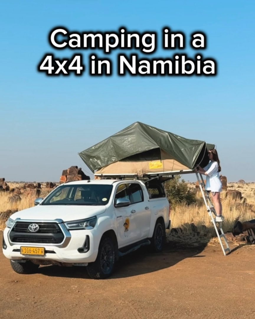 A woman climbing a ladder into a rooftop tent set up on a white 4x4 truck parked in a dry, rocky landscape, with bold text above reading “Camping in a 4x4 in Namibia.”