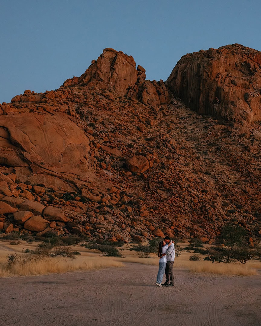 Couple standing and hugging in a dry, sandy desert in Spitzkoppe campsite with towering red rock cliffs in the background.