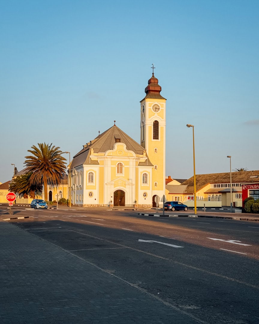 A light yellow and white church with a tall clock tower and red dome, bathed in warm sunset light on a quiet street with a palm tree and a few parked cars.