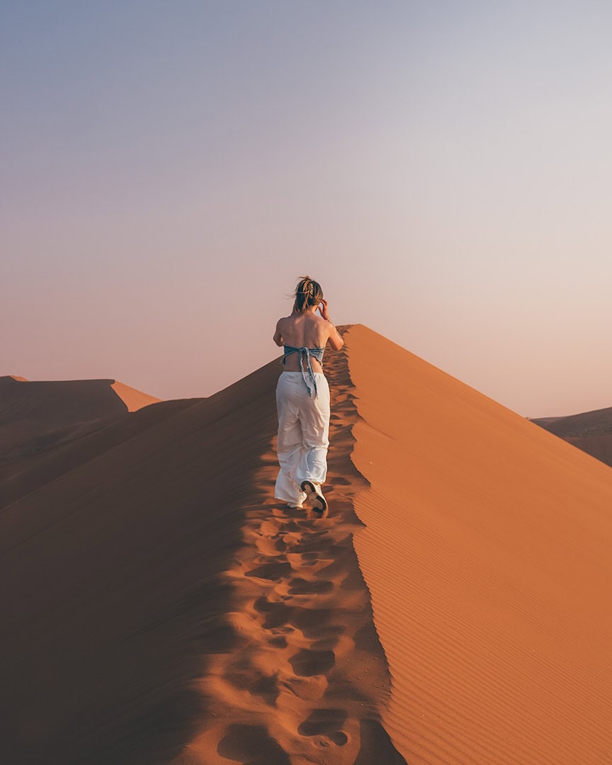 Woman walking along the ridge of a tall orange sand dune in Sossusvlei with footprints trailing behind her. The sky is clear and the desert stretches into the distance.