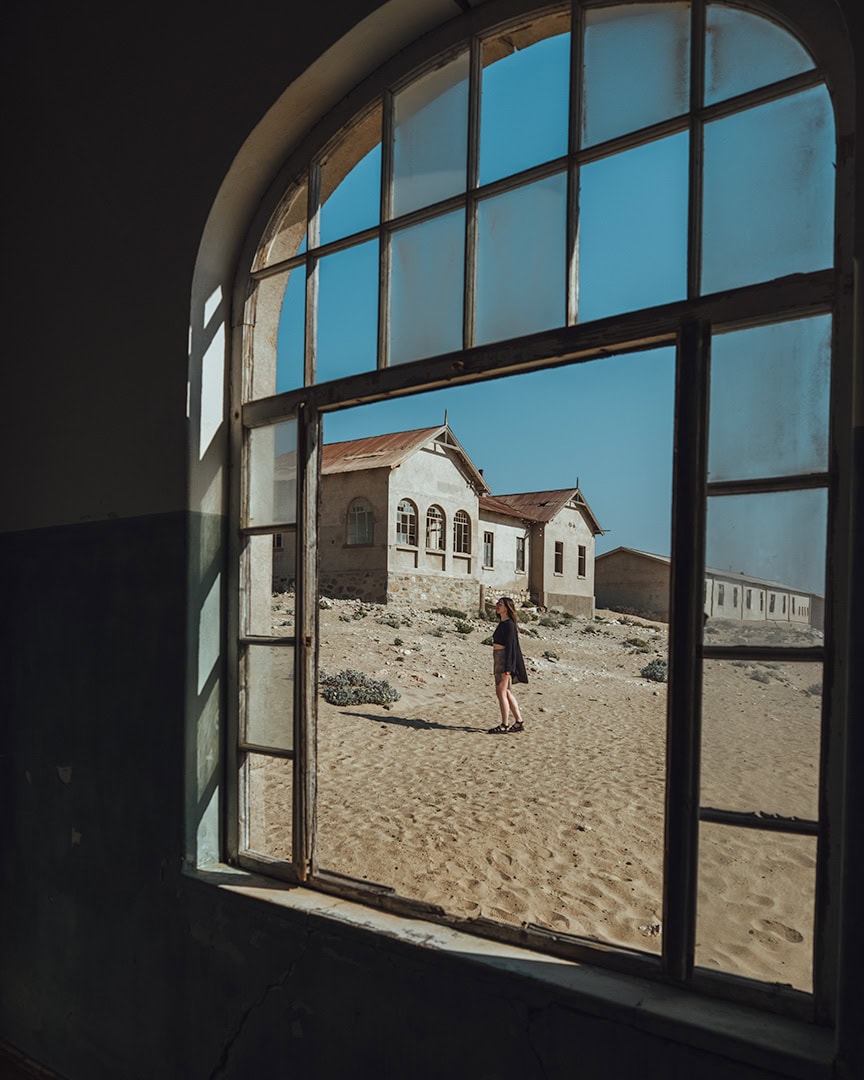 Woman walking across the sand-filled streets of Kolmanskop abandoned diamond mining ghost town, viewed through an arched hospital window, near Lüderitz, Namibia.