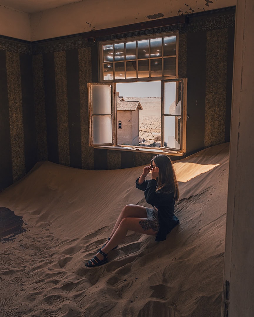 Woman sitting inside an abandoned room in Kolmanskop filled with sand dunes blowing in through an open window. Sunlight streams in and reveals the desert outside.