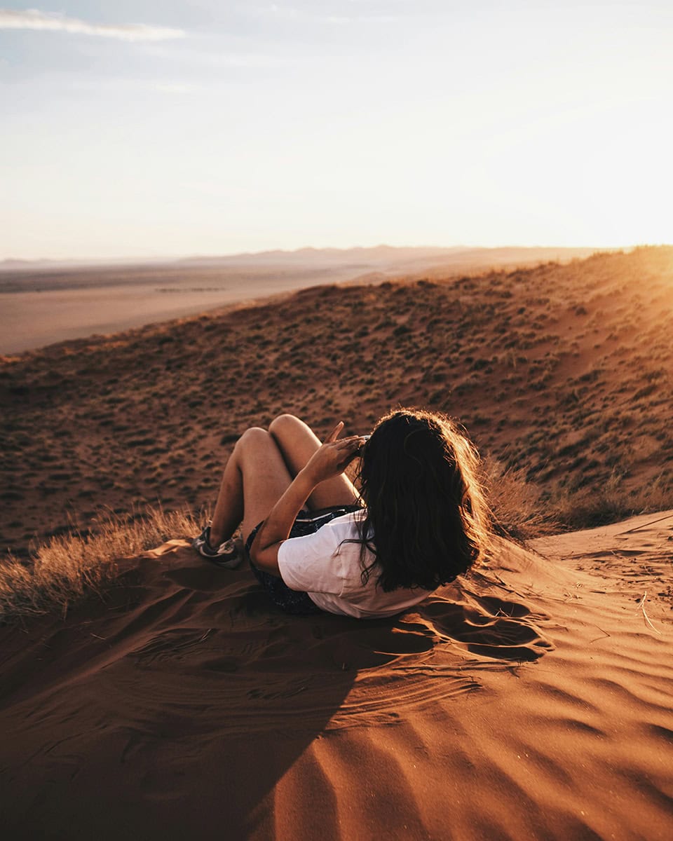 Woman lying on a sand dune at sunset overlooking a vast desert landscape.