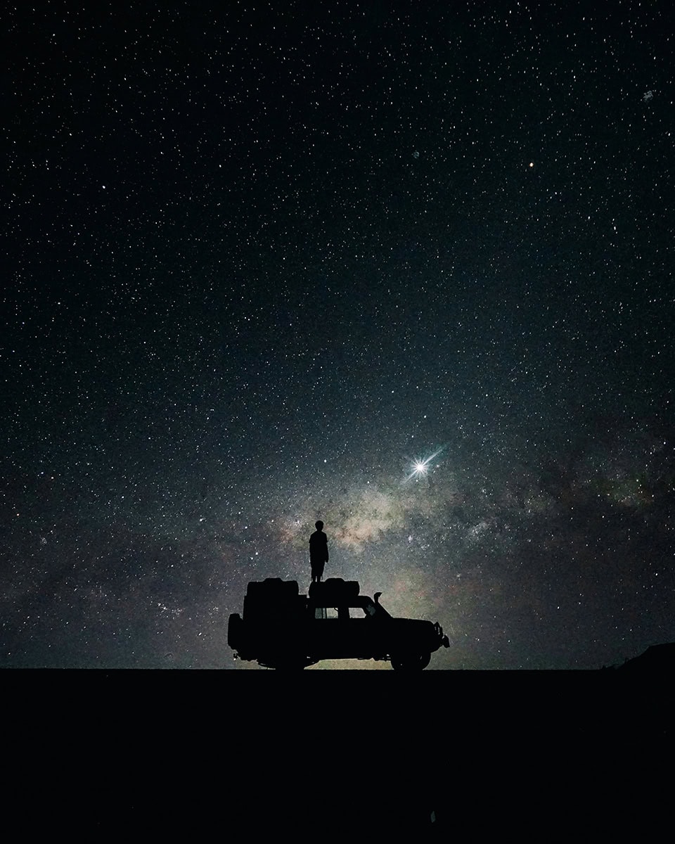 Silhouette of a person standing on top of a safari vehicle beneath a star-filled night sky and the Milky Way.