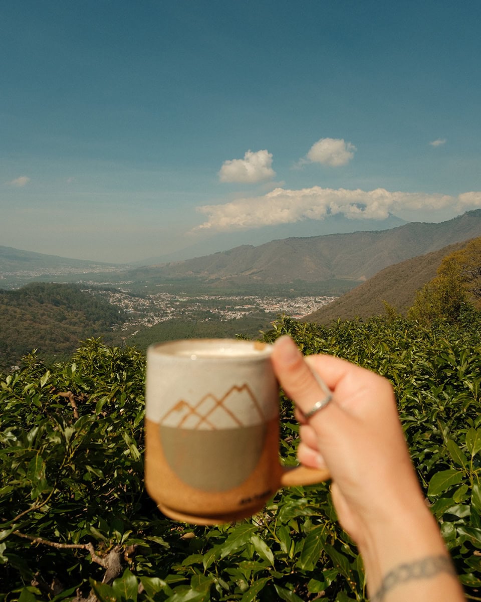 Hand holding a ceramic mug of coffee overlooking a green valley, mountains and volcanoes in the distance at Earth Lodge.