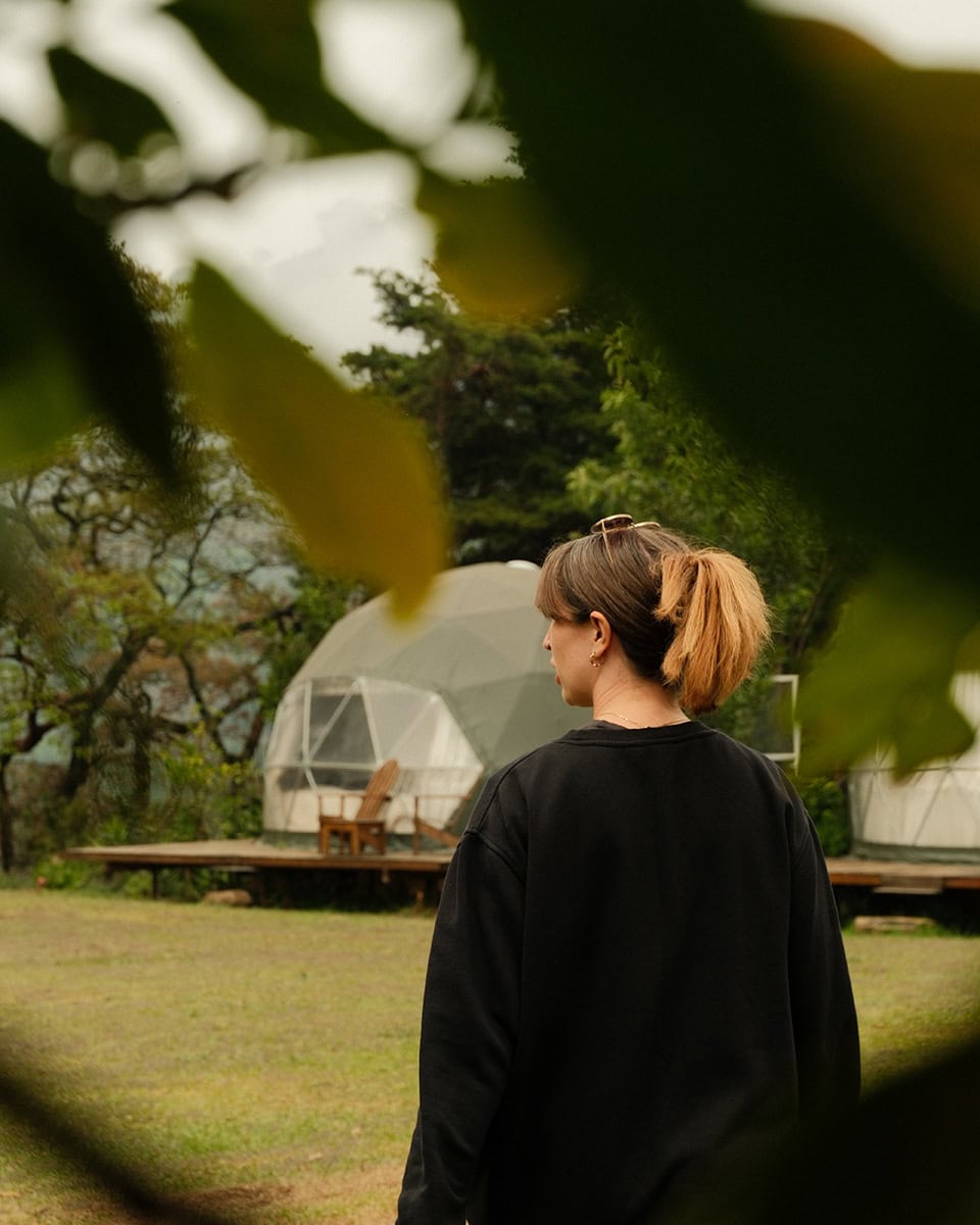 Woman seen from behind looking toward geodome tents at Earth Lodge framed by leaves in the foreground.