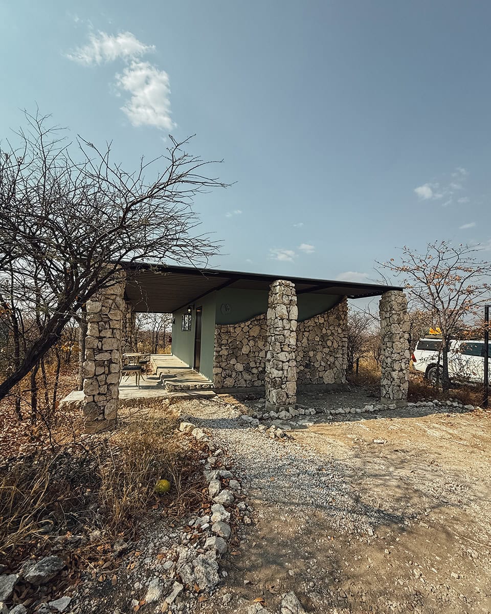 A small stone and concrete shelter with a covered porch sits in a dry bush landscape, with a vehicle partially visible nearby.