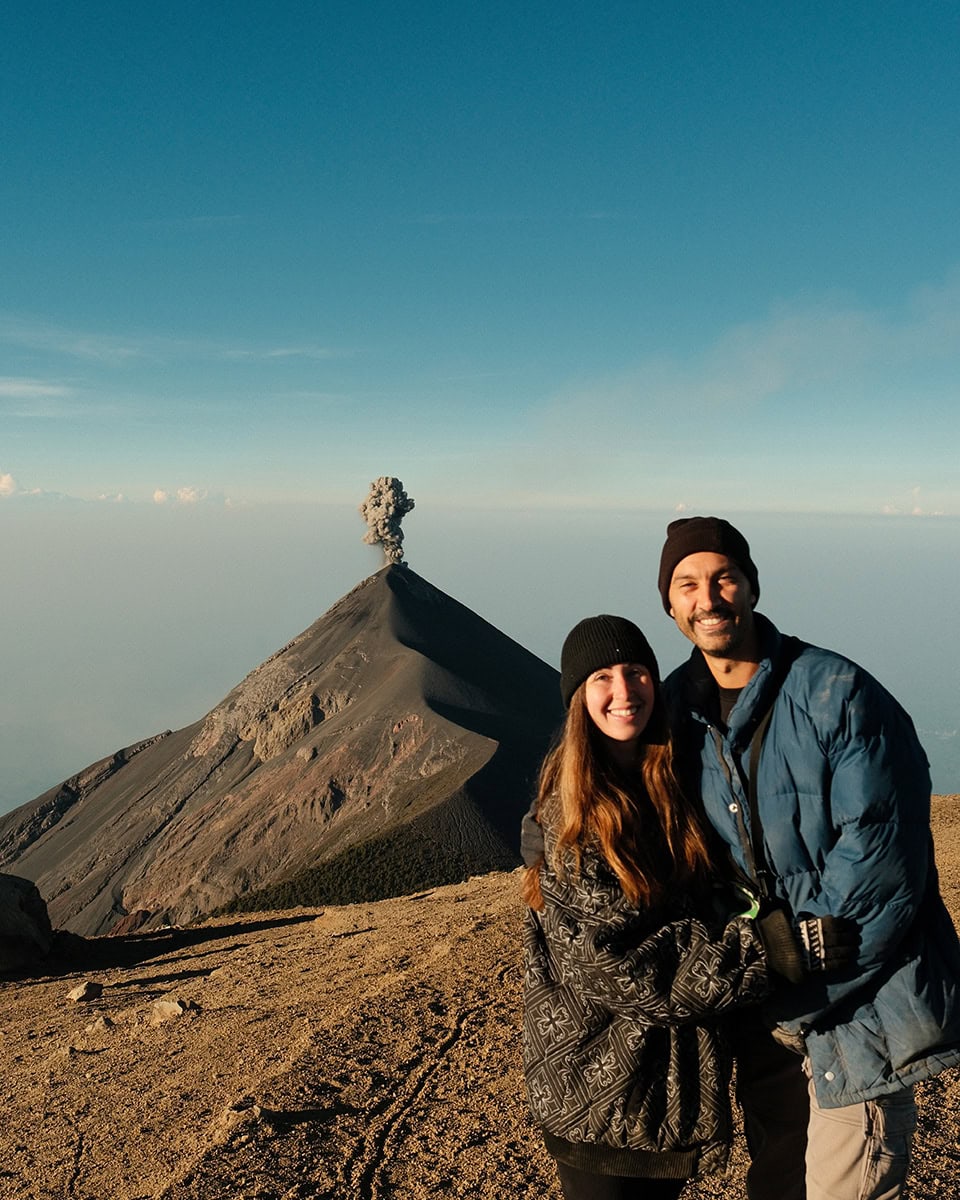 Couple posing together in front of Volcán de Fuego smoking in the background. The volcanic ridge rises sharply behind them under a bright blue morning sky.