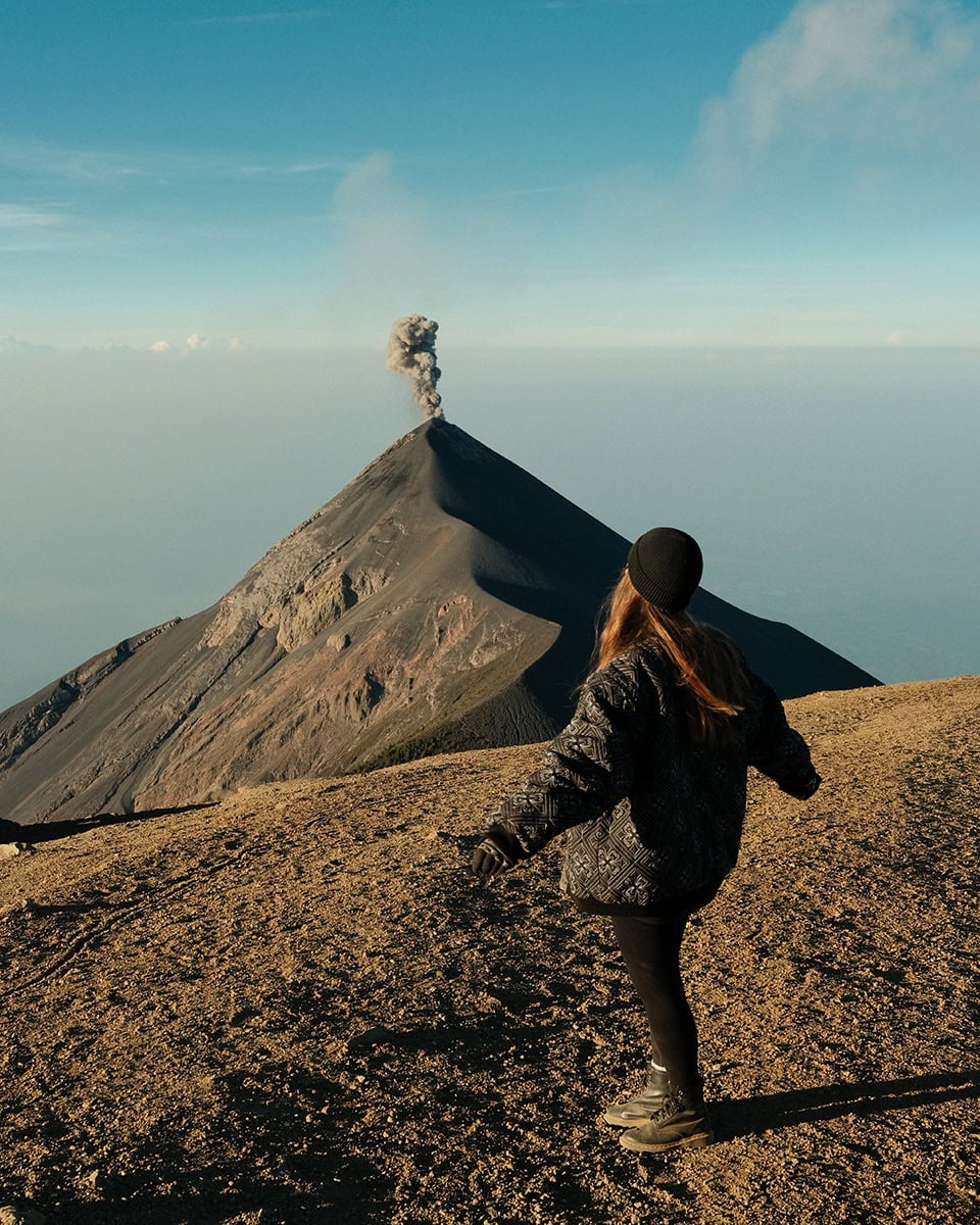 Woman walking along the Acatenango summit while looking toward Volcán de Fuego releasing smoke from the crater. The rocky volcanic terrain stretches above a sea of clouds.