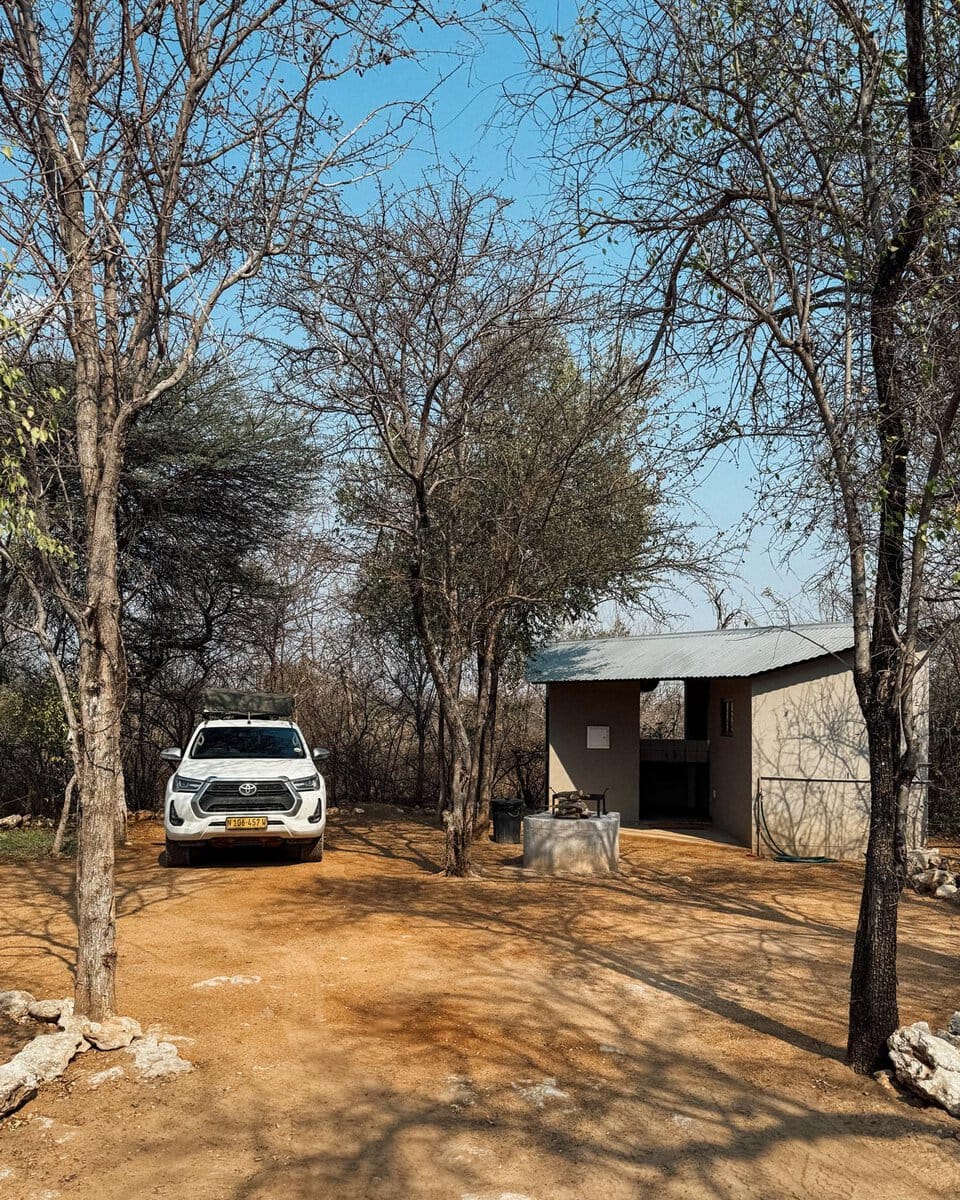 White 4x4 with a rooftop tent parked under trees next to a private shower block at Onguma Tamboti campsite near Etosha