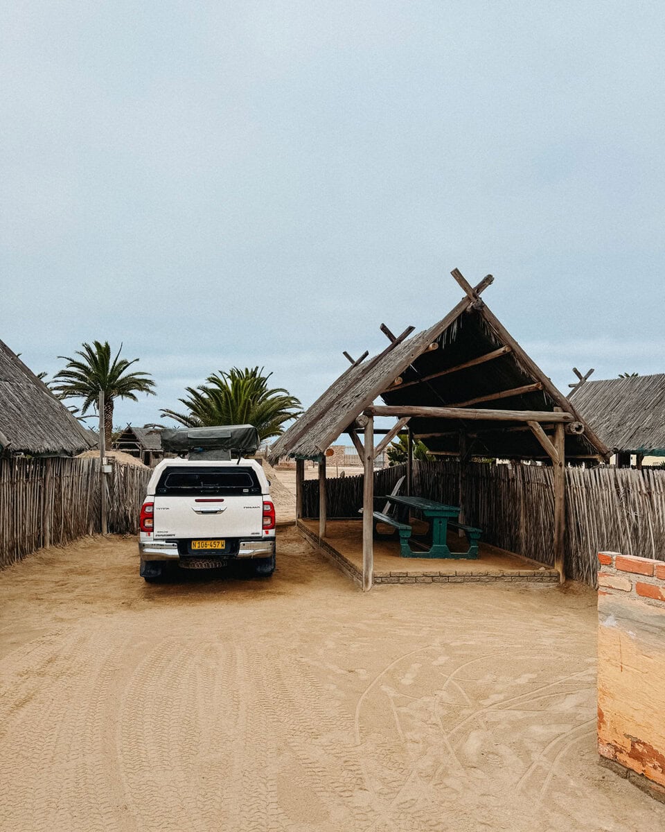 a white 4x4 parked beside a thatched picnic shelter at a sandy Tiger Reef campsite in Swakopmund
